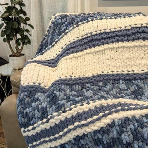 May include: A chunky knit throw blanket featuring thick, soft yarn in alternating horizontal stripes of solid cream white and a variegated blend of dark navy, medium blue, and light sky blue tones, showcasing a prominent, textured loop stitch pattern.