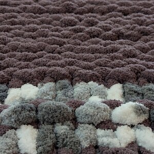 May include: A close-up of a handmade, chunky knit blanket. The blanket features a textured design with alternating rows of dark purple, white, and gray yarn. The soft, plush material creates a cozy and inviting aesthetic.