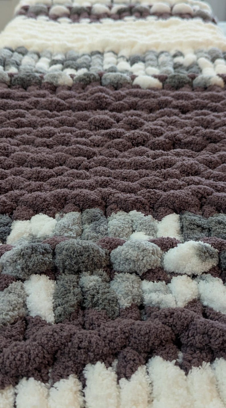 May include: A close-up of a handmade, chunky knit blanket. The blanket features a textured design with alternating rows of dark purple, white, and gray yarn. The soft, plush material creates a cozy and inviting aesthetic.