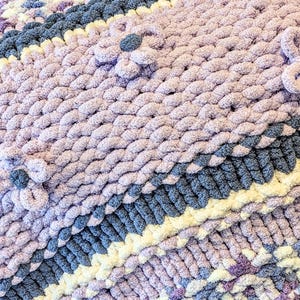 May include: A handmade, chunky knit blanket in shades of lavender, gray, and cream. The blanket features a textured design with raised floral accents. The soft, plush material is ideal for adding warmth and comfort to any space.