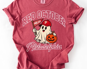 May include: A crimson short-sleeve t-shirt featuring a graphic of a ghost wearing a red baseball cap with a white P, holding a red pennant that says Phillies, and a jack-o'-lantern. The text reads RED OCTOBER in an arch and Philadelphia below.