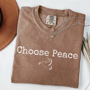 May include: A brown Comfort Colors t-shirt with the words "Choose Peace" and a dove graphic. A silver necklace with a small pendant is draped across the shirt. A brown hat is partially visible in the upper left corner.
