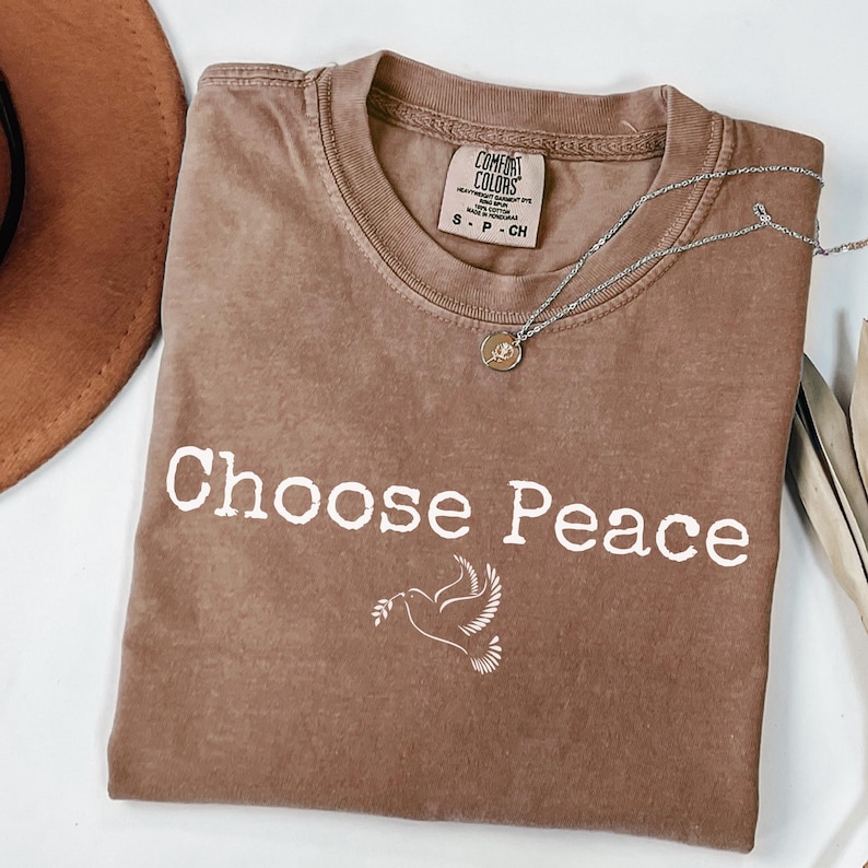 May include: A brown Comfort Colors t-shirt with the words "Choose Peace" and a dove graphic. A silver necklace with a small pendant is draped across the shirt. A brown hat is partially visible in the upper left corner.