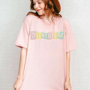 Puede incluir: Camiseta rosa claro con la palabra "Disneyland" impresa en colores pastel en el pecho. El texto est&aacute; delineado en dorado. La camiseta tiene mangas cortas y cuello redondo.