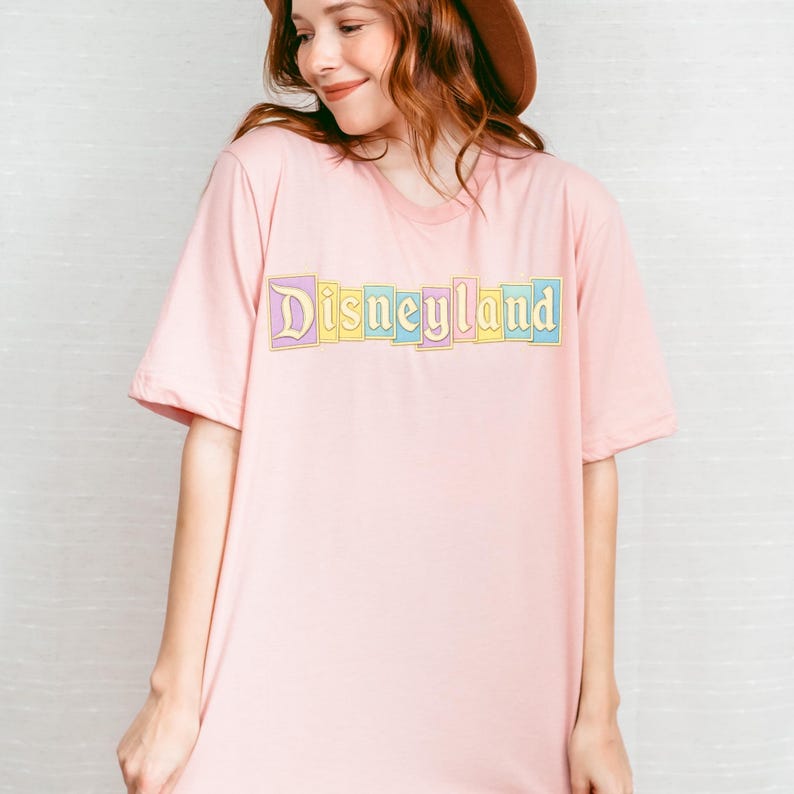 Puede incluir: Camiseta rosa claro con la palabra "Disneyland" impresa en colores pastel en el pecho. El texto est&aacute; delineado en dorado. La camiseta tiene mangas cortas y cuello redondo.