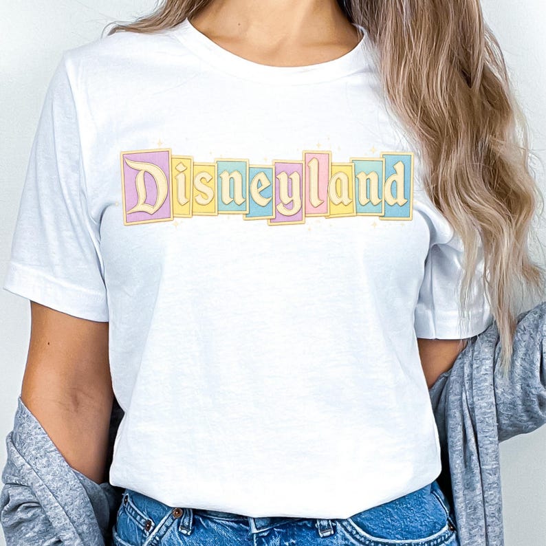 Puede incluir: Camiseta blanca con la palabra "Disneyland" en colores pastel. El texto est&aacute; en un dise&ntilde;o rectangular con una combinaci&oacute;n de colores azul claro, amarillo y rosa. La camiseta es de cuello redondo.