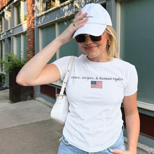 May include: White t-shirt with the text "stars, stripes, & human rights" and an American flag graphic. The person is wearing a white baseball cap and sunglasses.