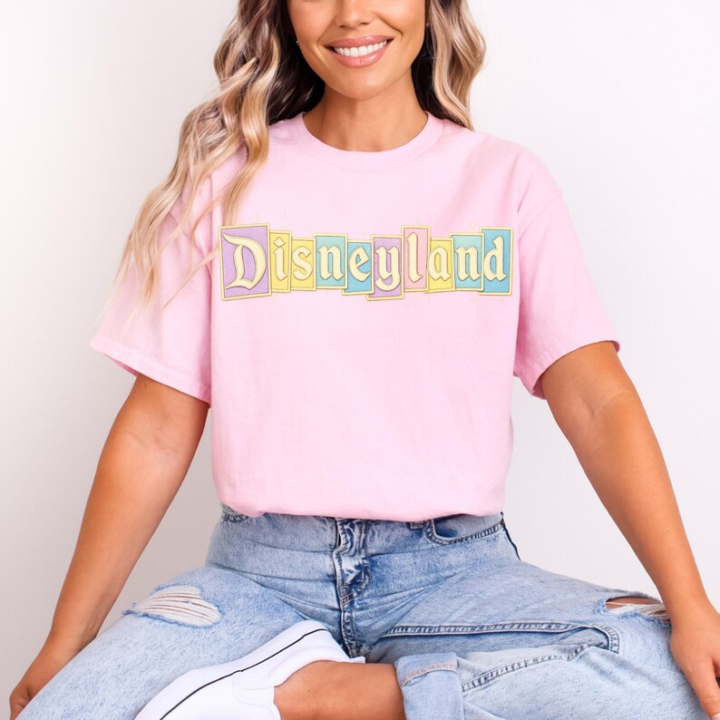 Puede incluir: Una camiseta rosa claro con la palabra "Disneyland" impresa en el pecho en colores pastel. El texto est&aacute; en un dise&ntilde;o de bloque rectangular. La camiseta es de manga corta y se lleva con vaqueros azules.