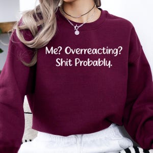 May include: A maroon long-sleeve sweatshirt featuring the white text Me? Overreacting? Shit Probably. The garment has a crew neckline and is worn with a silver-toned chain necklace featuring a circular pendant and a thin black choker necklace.