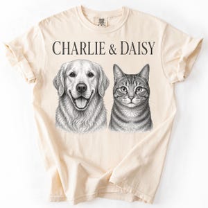 May include: A cream-colored short-sleeve t-shirt featuring a black ink-style illustration of a golden retriever and a tabby cat. The text CHARLIE & DAISY is printed in a serif font above the animal portraits on the front of the garment.