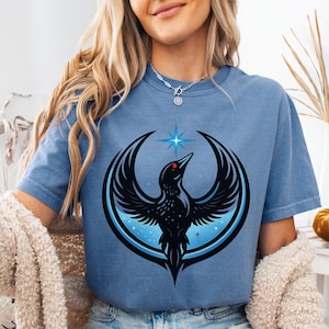 May include: A blue t-shirt featuring a stylized black bird with outstretched wings, a red eye, and a blue star above its head. The bird is set against a blue and black circular design. The shirt is a solid blue color.