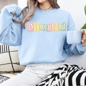 Puede incluir: Sudadera azul claro con la palabra "Disneyland" en una fuente colorida de estilo retro. El texto est&aacute; enmarcado por colores pastel. La sudadera tiene mangas largas y cuello redondo.
