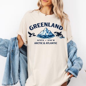 May include: Cream-colored t-shirt with a Greenland graphic. The design includes the word "GREENLAND" above an iceberg, a puffin, and a killer whale. Below the graphic are coordinates and the text "ARCTIC & ATLANTIC".