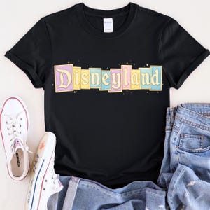 Puede incluir: Camiseta negra con la palabra "Disneyland" en colores pastel. El texto est&aacute; dispuesto en bloques rectangulares individuales con un dise&ntilde;o de estrellas. Zapatillas Converse blancas y vaqueros azules tambi&eacute;n est&aacute;n en la imagen.