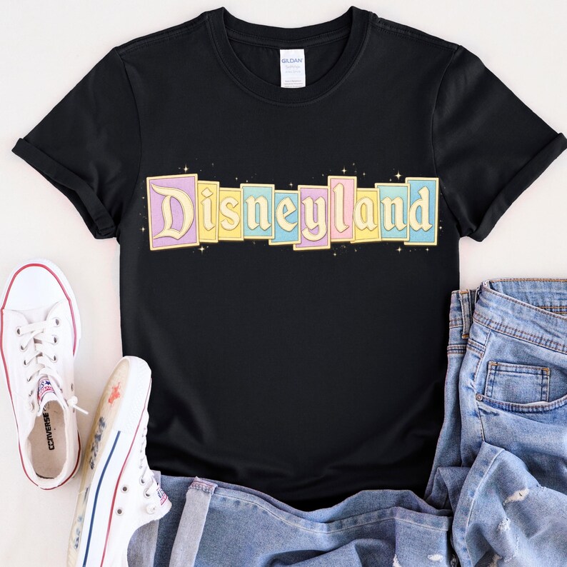 Puede incluir: Camiseta negra con la palabra "Disneyland" en colores pastel. El texto est&aacute; dispuesto en bloques rectangulares individuales con un dise&ntilde;o de estrellas. Zapatillas Converse blancas y vaqueros azules tambi&eacute;n est&aacute;n en la imagen.