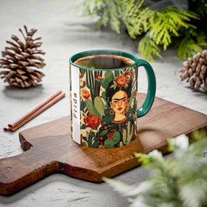 Może przedstawiać: Ceramiczny kubek z portretem Fridy Kahlo otoczonej kaktusami i kwiatami. Kubek ma zielony uchwyt i wnętrze, z napisem "Frida" wydrukowanym pionowo z boku. Kubek jest wypełniony ciemnym płynem.