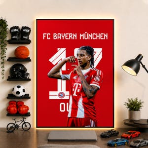 以下が含まれることがあります： 赤い背景に「FC BAYERN MÜNCHEN」の文字と大きな数字の17が描かれた長方形の壁掛けポスター。赤と白のユニフォームを着たサッカー選手が中央に配置され、手でジェスチャーをしている。ポスターは棚のある部屋に飾られている。
