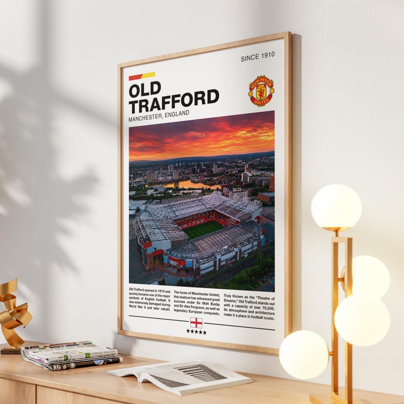 May include: A vertical rectangular poster in a light wood frame featuring a sunset aerial view of Old Trafford stadium in Manchester, England. The text includes the stadium name, the Manchester United crest, and historical facts about the venue.