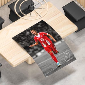 May include: A rectangular poster featuring a color photograph of a soccer player in a red Bayern Munich jersey with the number 9, running on a field. The background is in black and white, and a white stylized signature is visible in the corner.