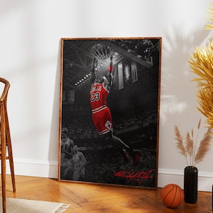 以下が含まれることがあります： 赤いユニフォームを着たバスケットボール選手が、空中でボールをダンクしようとしているフレーム入りのプリント。画像は主に白黒で、選手のユニフォームとバスケットボールのゴールは赤色です。選手の背番号は23です。