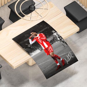 May include: A rectangular poster featuring a soccer player in a red and white jersey with the number 17, performing a goal celebration on a field. The background is black and white, and a white signature is visible in the bottom right corner.