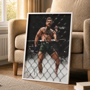 以下が含まれることがあります： 格闘家の白黒写真が額装されています。アスリートはUFCのロゴが入った緑色のショーツと黒い手袋を着用しています。フレームは白で、画像にはサインが入っています。