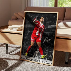 May include: A framed vertical poster featuring a color photograph of a soccer player in a red and white uniform celebrating a goal against a black and white stadium background. The player wears red shorts and a red jersey with white accents.