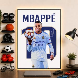 May include: A framed poster featuring a portrait of Kylian Mbappé in a white Real Madrid soccer jersey, holding a soccer ball. The text MBAPPÉ is printed at the top, with a large number 10 and a signature graphic in the background.