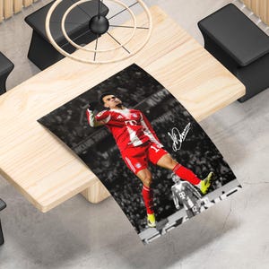 May include: A rectangular poster featuring a soccer player in a red and white jersey and shorts celebrating a goal. The background is a black and white stadium crowd scene. A white signature is printed on the right side of the image.