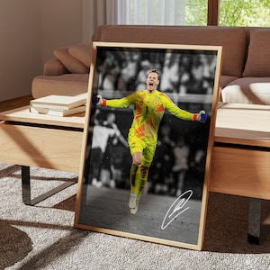 May include: A framed vertical poster featuring a color photograph of a soccer goalkeeper in a bright yellow and orange jersey with the number 1, celebrating with arms outstretched against a black and white blurred stadium background with a white signature.