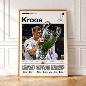 Puede incluir: Un póster enmarcado con marco dorado que presenta a Toni Kroos besando un trofeo. El póster incluye el nombre "Kroos", una cita y una biografía. El póster se exhibe en una pared blanca. Un artículo con temática de fútbol.