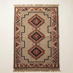 Puede incluir: Una alfombra rectangular con una base de color marrón claro y tres motivos centrales en forma de diamante en negro, naranja y marrón. La alfombra presenta un patrón geométrico en el borde y flecos en los extremos superior e inferior.
