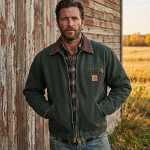 Könnte beinhalten: Dunkelgrüne Arbeitsjacke mit braunem Cordkragen und braunem Lederaufnäher auf der Brust. Die Jacke ist mit einem Reißverschluss geschlossen und wird über einem karierten Hemd und einer Jeans getragen. Der Hintergrund ist eine verwitterte Holzstruktur und ein Feld.