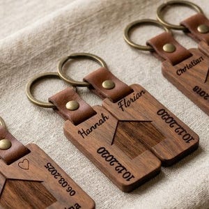Op de afbeelding: Drie paar rechthoekige houten sleutelhangers met bruine leren riempjes en messing ringen. Elk paar is voorzien van een gegraveerde huisomtrek, namen, datums en symbolen zoals hartjes of oneindigheidstekens met een donkere houtnerf.
