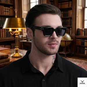 May include: Black square-framed sunglasses with a dark tint. The sunglasses have a thick frame and are worn by a person. The person is wearing a black collared shirt. The background features a library with bookshelves and a desk lamp.
