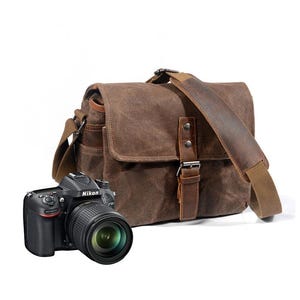 Puede incluir: Una bolsa para cámara de lona marrón con solapa de cuero, cierre de hebilla y una correa ancha para el hombro, colocada detrás de una cámara réflex digital Nikon D7100 negra con lente. La bolsa tiene costuras y detalles de metal visibles.