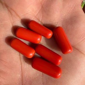 May include: Six bright red, cylindrical beads are displayed. The beads are smooth and have rounded ends. The beads are arranged in a scattered pattern, with some overlapping. The image is taken in natural light, highlighting the beads' color and texture.