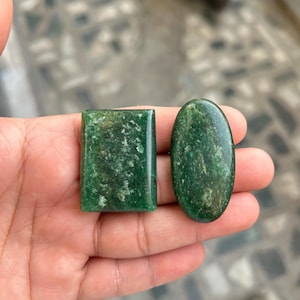 Puede incluir: Dos piedras preciosas verdes, una rectangular y otra ovalada, se sostienen en una mano. Las piedras preciosas tienen un aspecto moteado y una superficie pulida. La piedra rectangular es de corte cuadrado, mientras que la piedra ovalada es lisa y redondeada. Las piedras son probablemente para la fabricación de joyas.