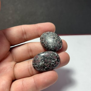 Puede incluir: Dos piedras pulidas de forma ovalada con un patrón moteado de negro, gris y rojo oscuro. Las piedras se sostienen en una mano, mostrando sus superficies lisas y reflectantes. Las piedras parecen ser piedras preciosas naturales.