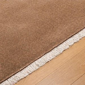 May include: A brown rug with a textured surface and a cream-colored fringe. The rug is placed on a light-colored wooden floor, creating a contrast in textures and colors. The rug's edge is visible.