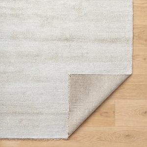 May include: A rectangular, off-white rug with a subtle, textured pattern. The corner is folded back, revealing a beige underside. The rug is placed on a light-colored wooden floor, showcasing its design and texture.