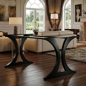 Può includere: Un tavolo consolle nero con base curva e piano in vetro. Il tavolo si trova in un soggiorno con camino, divani beige e pavimenti in legno. La stanza è ben illuminata.