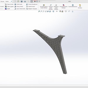 Könnte beinhalten: Ein stilisiertes graues Metall-Tischbein mit geschwungenem Design und einer Reihe kleiner, gleichmäßig verteilter kreisförmiger Aussparungen. Das Bein hat zwei flache Montageflächen oben und eine breitere Basis.