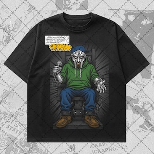 May include: A black short-sleeve t-shirt featuring a graphic of a figure in a metal mask, green hoodie, and blue jeans sitting in a chair. The graphic includes the text MF DOOM and a speech bubble with text about heroes and brutality.