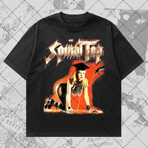 May include: Black t-shirt featuring the band name "Spinal Tap" in a metallic, gothic font. The graphic depicts a woman in a provocative pose, wearing a graduation cap and holding a whip. The background is a gradient of red and orange.