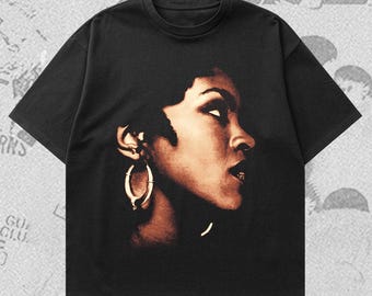 Puede incluir: Camiseta negra con un retrato en tonos sepia de una mujer de perfil, con grandes pendientes de aro. La imagen está centrada en la parte delantera de la camiseta. La mujer tiene el pelo oscuro y mira hacia arriba. La camiseta tiene mangas cortas y cuello redondo.