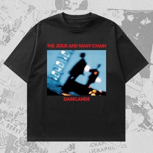 Op de afbeelding: Zwart T-shirt met de bandnaam "THE JESUS AND MARY CHAIN" in rode letters boven een wazige afbeelding. Het woord "DARKLANDS" staat eronder in het rood. Het T-shirt heeft een ronde hals.