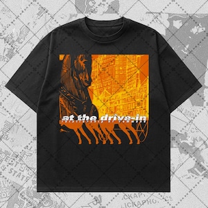 May include: A black short-sleeve t-shirt featuring a graphic print in shades of orange and yellow. The design depicts a horse-drawn carriage in front of a city building, with the text "at the drive-in" and "relationship of command" printed.