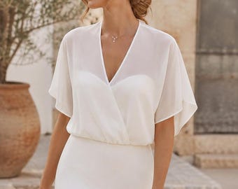 Pode incluir: Um vestido branco com uma sobreposição transparente de mangas curtas, decote em V e corpete estilo envelope. A peça possui uma cintura ajustada e uma saia lisa de cor sólida que se estende para baixo, criando uma silhueta limpa e estruturada.