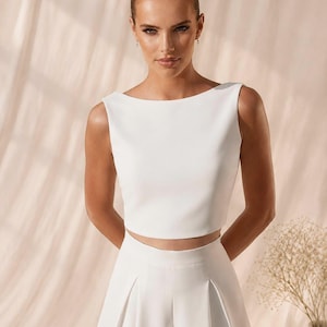 Könnte beinhalten: Ein weißes ärmelloses Crop-Top und eine hoch taillierte Hose. Das Oberteil hat einen U-Boot-Ausschnitt und die Hose hat Falten. Das Outfit besteht aus einem glatten, blickdichten Stoff. Das Model trägt das Outfit vor einem neutralen Hintergrund.